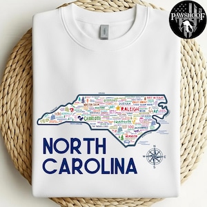 Puede incluir: Sudadera blanca con un mapa detallado de Carolina del Norte, lleno de nombres de ciudades y puntos de referencia. Las palabras "NORTH CAROLINA" est&aacute;n impresas en azul marino debajo. Tambi&eacute;n se incluye una rosa de los vientos.