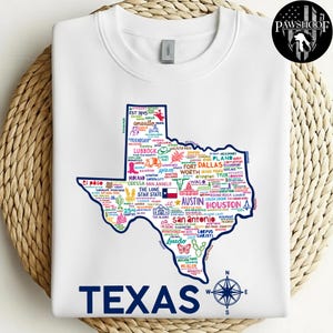 May include: White crewneck sweatshirt featuring a detailed map of Texas, filled with city names and icons. The word "TEXAS" is printed in navy blue at the bottom, with a compass rose graphic. A Pawshoof logo is in the upper right corner.