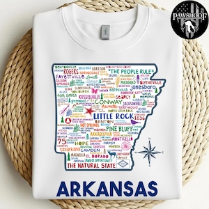 Puede incluir: Sudadera blanca de cuello redondo con un mapa colorido de Arkansas con nombres de ciudades y puntos de referencia. El texto "ARKANSAS" se muestra en letras azules grandes en la parte inferior. Una rosa de los vientos está en el lado derecho. La camisa también tiene el logo de Pawshoof.