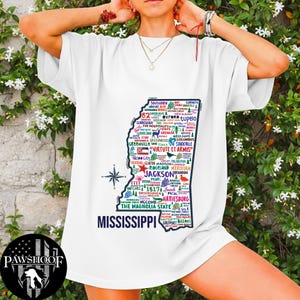 May include: White t-shirt featuring a colorful map of Mississippi, filled with city names and the state motto "Virtute et Armis." The text "Mississippi" is printed below the map. The shirt also has a compass rose design.