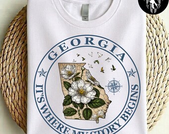 Może przedstawiać: Biały t-shirt z niebieską grafiką w kształcie koła i napisem GEORGIA IT'S WHERE MY STORY BEGINS. Wzór zawiera mapę stanu Georgia wypełnioną białymi kwiatami róży Cherokee, zielonymi liśćmi, różą wiatrów oraz małymi latającymi ptakami.