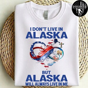 May include: A white t-shirt featuring the blue text I DON'T LIVE IN ALASKA BUT ALASKA WILL ALWAYS LIVE IN ME. The design includes an infinity symbol shaped like a heart with American flag patterns, a bear, a crab, a whale, and a totem pole.