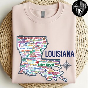 May include: A light pink sweatshirt featuring a detailed map of Louisiana, with city names and the state's outline in navy blue. The text "UNION, JUSTICE, AND CONFIDENCE" is at the top. A compass rose is at the bottom right.