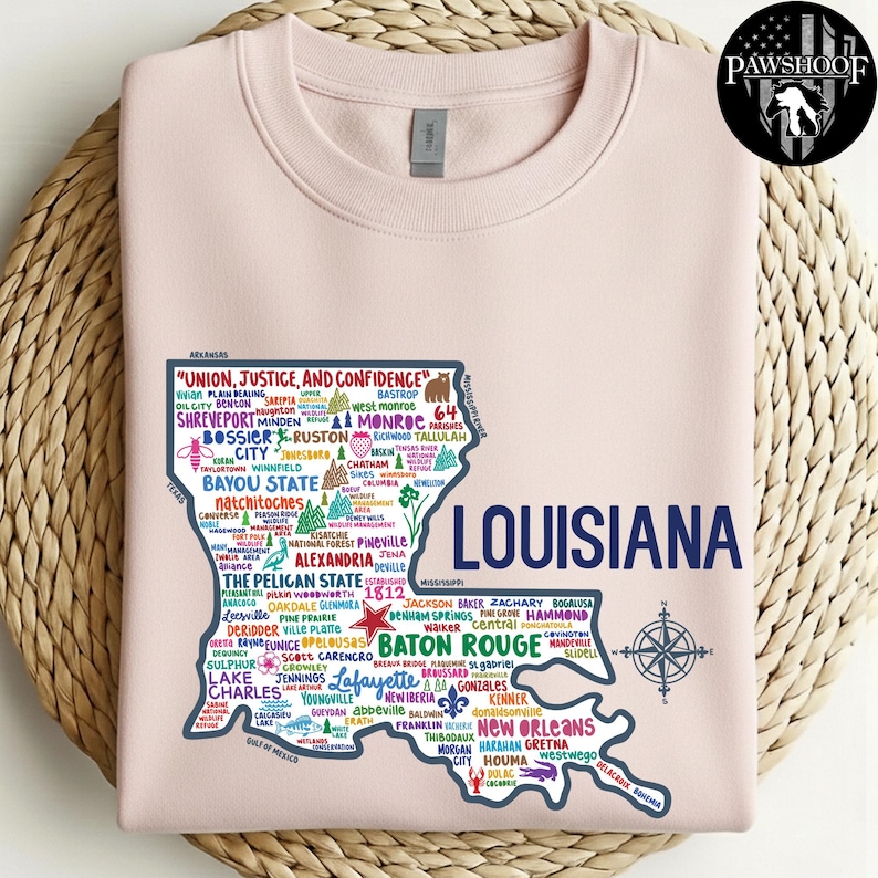 May include: A light pink sweatshirt featuring a detailed map of Louisiana, with city names and the state's outline in navy blue. The text "UNION, JUSTICE, AND CONFIDENCE" is at the top. A compass rose is at the bottom right.
