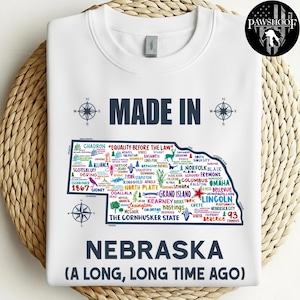 Op de afbeelding: Witte sweatshirt met een kaart van Nebraska, met stadsnamen en de tekst "MADE IN NEBRASKA (A LONG, LONG TIME AGO)". Bevat kompasroosafbeeldingen en de zin "Equality Before The Law".