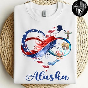 May include: White sweatshirt with a heart-shaped design and the word "Alaska" in blue script. The heart design includes elements of the American flag, a feather, and Alaskan imagery such as a bear, whale, and totem pole.