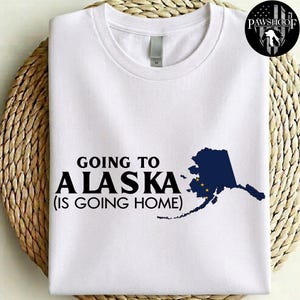 以下が含まれることがあります： 黒い文字で「GOING TO ALASKA (IS GOING HOME)」とプリントされ、北斗七星と北極星を表す黄色い星が描かれたアラスカ州の濃い青色のシルエット地図がフロントにデザインされた、白い半袖のクルーネックTシャツ。