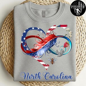 May include: A heather gray crewneck sweatshirt featuring a graphic of an infinity heart symbol colored with red, white, and blue American flag patterns. The design includes a North Carolina state map, a strawberry, a squirrel, and a bee.
