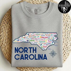 Puede incluir: Una sudadera gris con un contorno detallado de Carolina del Norte, llena de nombres de ciudades e iconos. El texto "NORTH CAROLINA" est&aacute; impreso en azul marino debajo del mapa. Una rosa de los vientos est&aacute; a la derecha.
