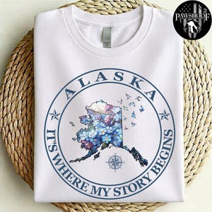 May include: A white short-sleeved t-shirt featuring a circular blue graphic. Inside the circle is a map of Alaska filled with blue and purple floral patterns, small flying birds, and a compass rose. The text reads ALASKA IT'S WHERE MY STORY BEGINS.