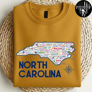 Puede incluir: Sudadera de color mostaza con un contorno detallado de Carolina del Norte, llena de nombres de ciudades y puntos de referencia. Las palabras "NORTH CAROLINA" est&aacute;n impresas en azul marino debajo del mapa. Una rosa de los vientos est&aacute; en la parte inferior derecha.