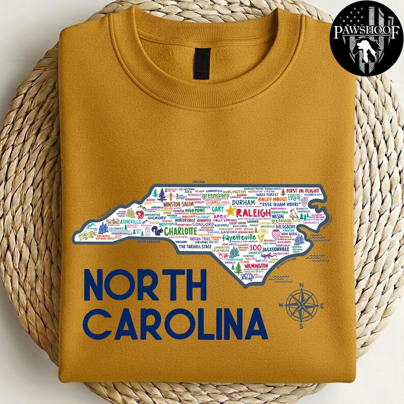 Puede incluir: Sudadera de color mostaza con un contorno detallado de Carolina del Norte, llena de nombres de ciudades y puntos de referencia. Las palabras "NORTH CAROLINA" est&aacute;n impresas en azul marino debajo del mapa. Una rosa de los vientos est&aacute; en la parte inferior derecha.
