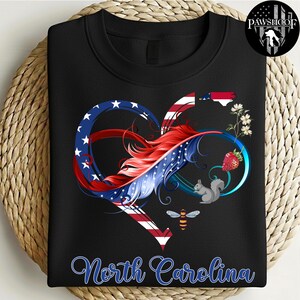 May include: A black crewneck sweatshirt featuring a graphic of an infinity heart shape colored with the American flag pattern. The design includes a red feather, a North Carolina state outline, a strawberry, a squirrel, a bee, and flowers.