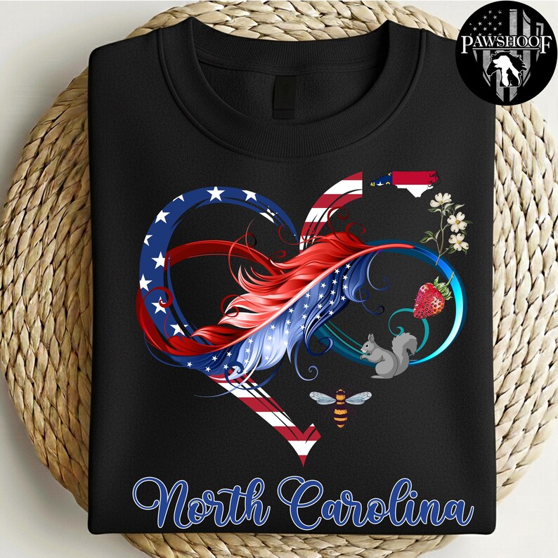 May include: A black crewneck sweatshirt featuring a graphic of an infinity heart shape colored with the American flag pattern. The design includes a red feather, a North Carolina state outline, a strawberry, a squirrel, a bee, and flowers.