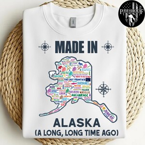 May include: White sweatshirt with a map of Alaska, featuring city names and landmarks. The text "MADE IN ALASKA (A LONG, LONG TIME AGO)" is printed on the front, along with compass rose designs.