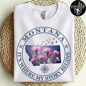 May include: A white short-sleeved t-shirt featuring a circular graphic with the text MONTANA IT'S WHERE MY STORY BEGINS. The design includes a map of Montana filled with colorful floral illustrations, small flying birds, and a compass rose below.