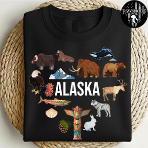 May include: A black crewneck sweatshirt featuring the word ALASKA in white text surrounded by illustrations of a muskox, walrus, orca, mammoth, brown bear, bald eagle, crab, salmon, seashell, caribou, wolf, rabbit, pinecone, and a colorful totem pole.