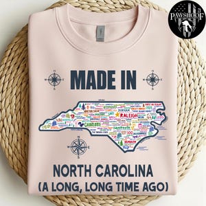 May include: A pale pink sweatshirt featuring the text "MADE IN" above a map of North Carolina, filled with city names and compass rose illustrations. The words "NORTH CAROLINA (A LONG, LONG TIME AGO)" are printed below the map.