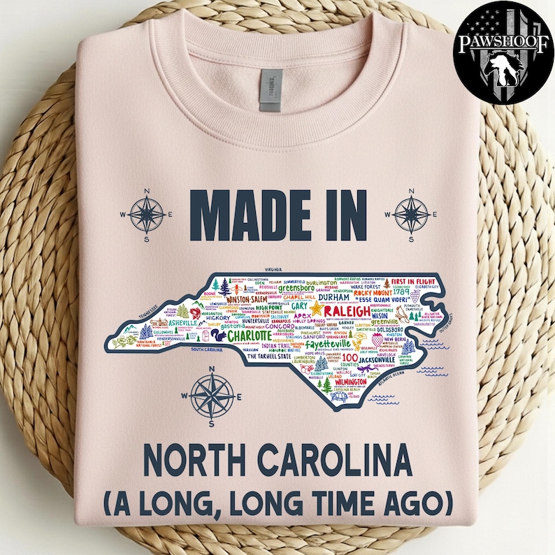 May include: A pale pink sweatshirt featuring the text "MADE IN" above a map of North Carolina, filled with city names and compass rose illustrations. The words "NORTH CAROLINA (A LONG, LONG TIME AGO)" are printed below the map.