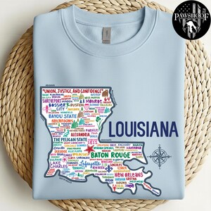 May include: Light blue sweatshirt featuring a map of Louisiana with city names and a compass rose. The state outline is filled with text, including "Union, Justice, and Confidence" and "The Pelican State". A PawShoof logo is in the upper right corner.