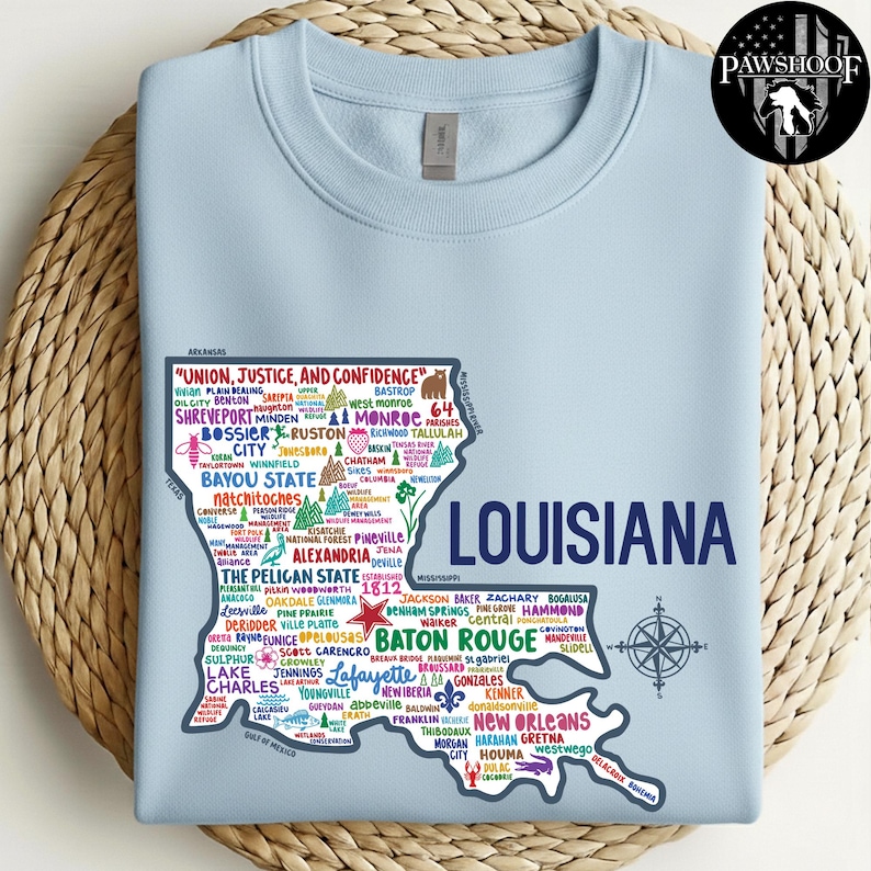 May include: Light blue sweatshirt featuring a map of Louisiana with city names and a compass rose. The state outline is filled with text, including "Union, Justice, and Confidence" and "The Pelican State". A PawShoof logo is in the upper right corner.