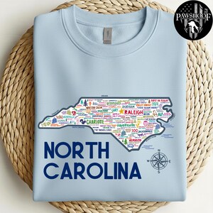 Puede incluir: Sudadera azul claro con un contorno detallado de Carolina del Norte lleno de nombres de ciudades y puntos de referencia. El texto "NORTH CAROLINA" est&aacute; impreso en azul marino debajo del mapa. Una rosa de los vientos est&aacute; en la parte inferior derecha.