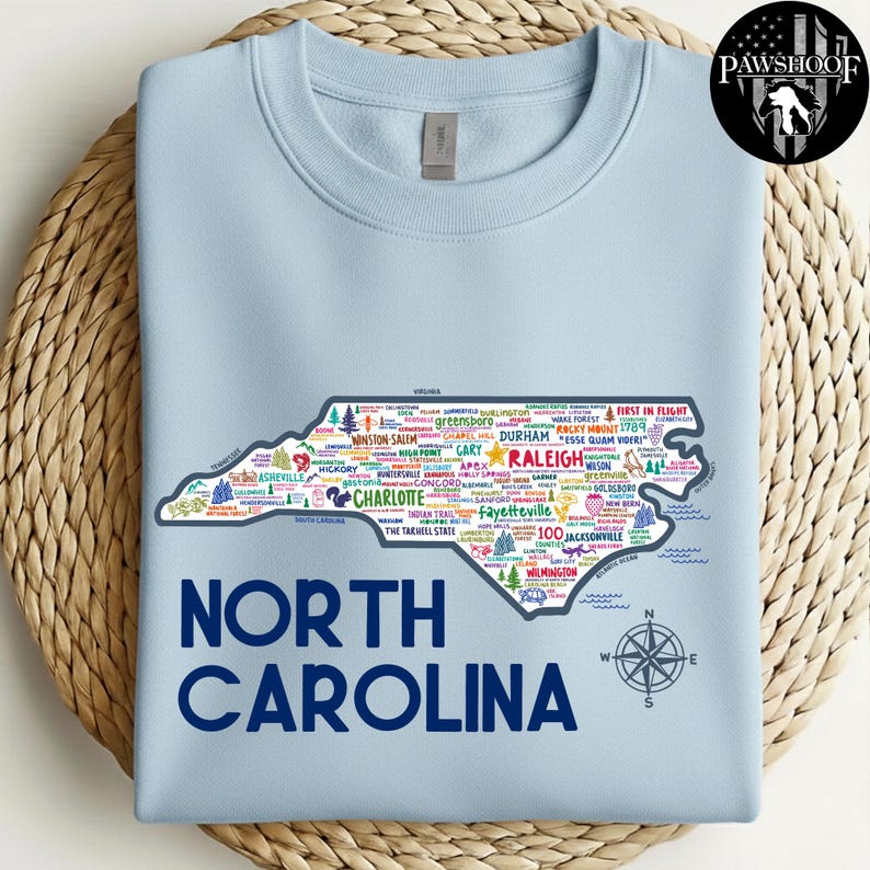 Puede incluir: Sudadera azul claro con un contorno detallado de Carolina del Norte lleno de nombres de ciudades y puntos de referencia. El texto "NORTH CAROLINA" est&aacute; impreso en azul marino debajo del mapa. Una rosa de los vientos est&aacute; en la parte inferior derecha.