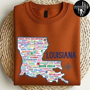 May include: Rust-colored sweatshirt featuring a map of Louisiana with city names and the text "LOUISIANA" in blue. The map includes the phrase "UNION, JUSTICE, AND CONFIDENCE". A compass rose graphic is on the right.
