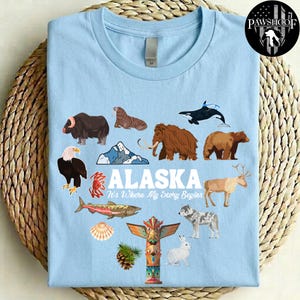 May include: A light blue t-shirt featuring a graphic print of various Alaskan wildlife including a muskox, walrus, orca, mammoth, bear, eagle, crab, salmon, caribou, wolf, and rabbit, along with a totem pole, mountain, and the text ALASKA It's Where My Story Begins.