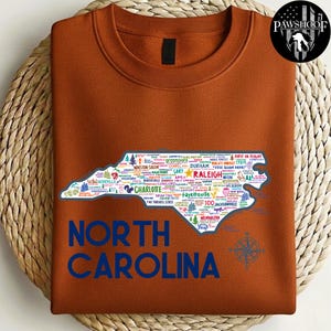 Puede incluir: Una sudadera de color &oacute;xido con un contorno detallado de Carolina del Norte lleno de nombres de ciudades e iconos. Las palabras "NORTH CAROLINA" est&aacute;n impresas en azul debajo del mapa. Tambi&eacute;n se ve una rosa de los vientos.