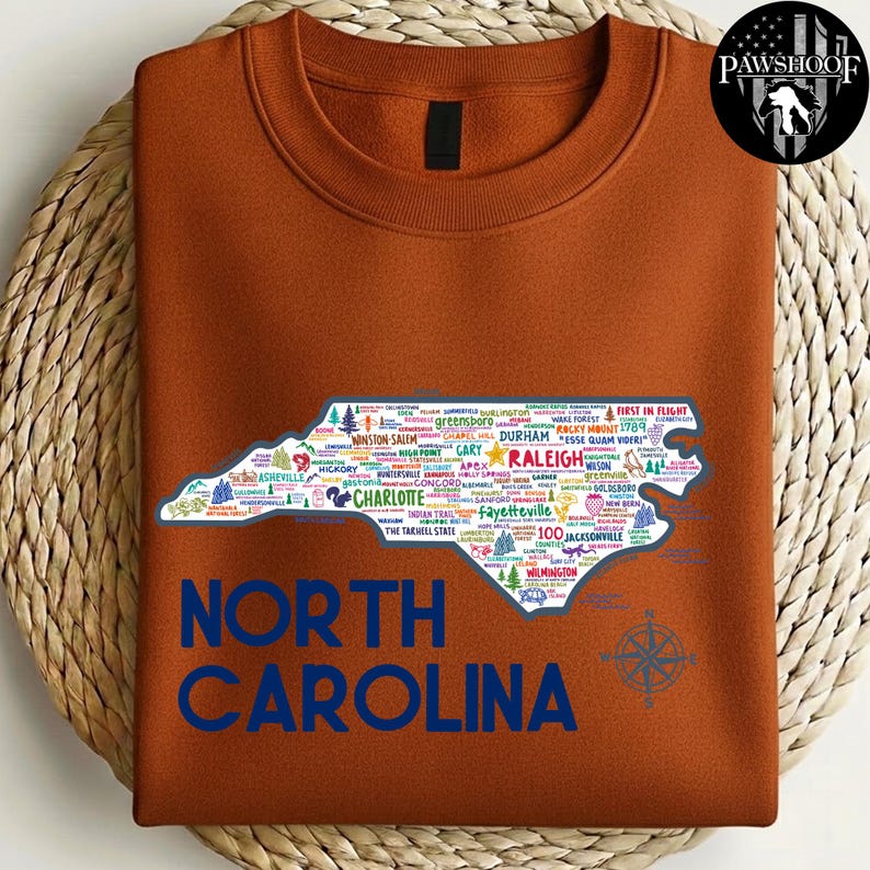 Puede incluir: Una sudadera de color &oacute;xido con un contorno detallado de Carolina del Norte lleno de nombres de ciudades e iconos. Las palabras "NORTH CAROLINA" est&aacute;n impresas en azul debajo del mapa. Tambi&eacute;n se ve una rosa de los vientos.