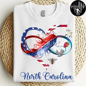 May include: A white sweatshirt featuring a graphic of an infinity heart shape with red, white, and blue patterns. The design includes a North Carolina state outline, a strawberry, a squirrel, a bee, and white flowers. The text North Carolina is below.