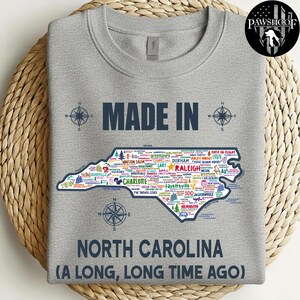 May include: A grey sweatshirt featuring the text "MADE IN NORTH CAROLINA (A LONG, LONG TIME AGO)". The design includes a map of North Carolina with city names and compass graphics.