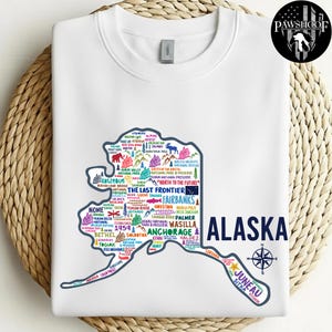 以下が含まれることがあります： アラスカの都市名とランドマークが描かれたカラフルな地図がプリントされた、白いクルーネックのスウェットシャツ。「ALASKA」の文字はネイビーブルーで印刷され、羅針盤のグラフィックが描かれています。スウェットシャツは丸襟で、ゆったりとしたフィット感です。