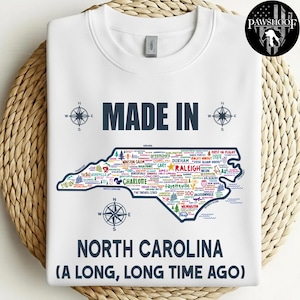May include: White sweatshirt with a map of North Carolina, featuring city names and the text "MADE IN NORTH CAROLINA (A LONG, LONG TIME AGO)". Compass rose designs are on the top and bottom. The sweatshirt has a crew neck.