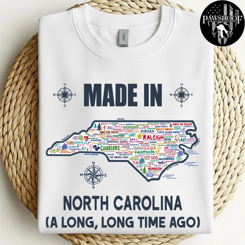 May include: White sweatshirt with a map of North Carolina, featuring city names and the text "MADE IN NORTH CAROLINA (A LONG, LONG TIME AGO)". Compass rose designs are on the top and bottom. The sweatshirt has a crew neck.