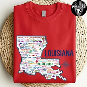 May include: A red sweatshirt featuring a detailed map of Louisiana, with city names and the state outline in white and blue. The text "LOUISIANA" is in large blue letters, and the state motto is at the top. A PawShoof logo is in the upper right corner.