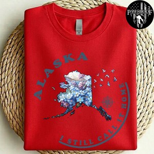 Puede incluir: Una camiseta roja de manga corta con un gráfico del estado de Alaska relleno de patrones florales azules y morados. El texto ALASKA e I STILL CALL IT HOME rodea el mapa, acompañado de pequeñas siluetas de aves y una brújula.
