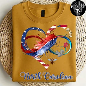 May include: A mustard yellow crewneck sweatshirt featuring a graphic of an infinity heart shape with red, white, and blue stripes and stars. The design includes a North Carolina state outline, a strawberry, a squirrel, a bee, and the text North Carolina.