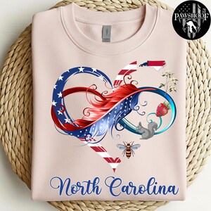 May include: A light pink crewneck sweatshirt featuring a graphic of an infinity heart symbol colored with red, white, and blue stars and stripes. The design includes a North Carolina state map, a strawberry, a squirrel, a bee, and flowers.