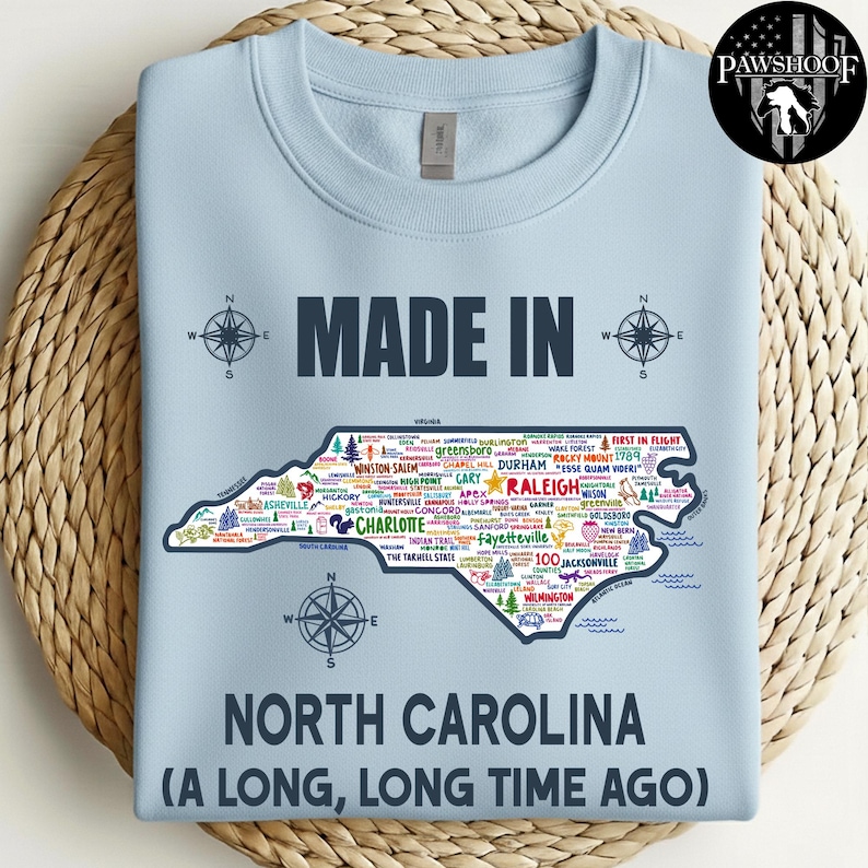 May include: Pale blue sweatshirt featuring a map of North Carolina with city names, the words "MADE IN" and "NORTH CAROLINA (A LONG, LONG TIME AGO)". The garment is a crew neck style, displayed on a woven surface.