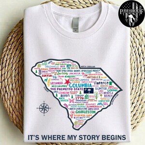 May include: A white short-sleeved t-shirt featuring a colorful graphic of the South Carolina state map filled with city names, icons, and the text 'THE PALMETTO STATE' and 'IT'S WHERE MY STORY BEGINS'. A compass rose is printed to the left of the map.