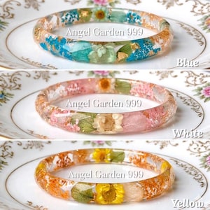 Op de afbeelding: Drie harsarmbanden met gedroogde bloemen en goudbladinsluitsels. De bovenste armband is blauw, de middelste roze en de onderste geel. Elke armband heeft de tekst "Angel Garden 999". De armbanden liggen op een wit bord.