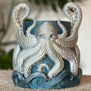 May include: A decorative planter featuring an octopus design. The octopus is silver with blue accents, including the tentacles and a wave-like base. The planter is cylindrical and suitable for indoor or outdoor use.