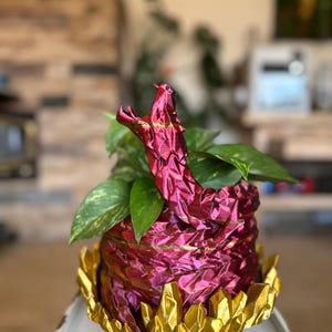 May include: A decorative planter with a red and gold snake design. The planter is filled with green leafy plants. The snake's head is open, as if it is hissing. The base of the planter is gold and has a crystal-like design.