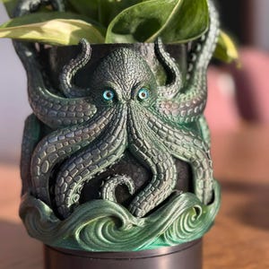 May include: A detailed, dark green and black octopus-themed planter pot with blue eyes. The tentacles wrap around the pot, and the base features a wave design. The pot holds green and yellow leafy plants.