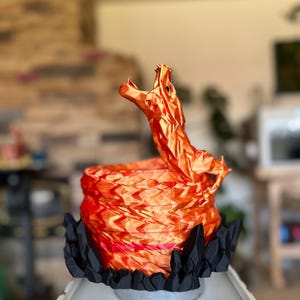 May include: A 3D-printed sculpture of a dragon in shades of orange and red, with a black base. The dragon is posed with its head raised and mouth open. The sculpture is on a white surface.