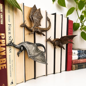 May include: Three dragon-shaped bookends in metallic bronze and silver finishes, each featuring detailed scales, wings, and horns. These decorative figures are positioned between vertical books, appearing to crawl across the spines of the volumes.