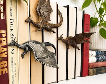 May include: Three dragon-shaped bookends in metallic bronze and silver finishes, each featuring detailed scales, wings, and horns. These decorative figures are positioned between vertical books, appearing to crawl across the spines of the volumes.