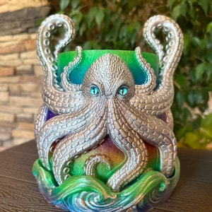 May include: A cylindrical container featuring a silver octopus with textured tentacles and bright blue eyes. The base of the container is decorated with sculpted waves, and the body displays a vibrant gradient of green, blue, and purple colors.
